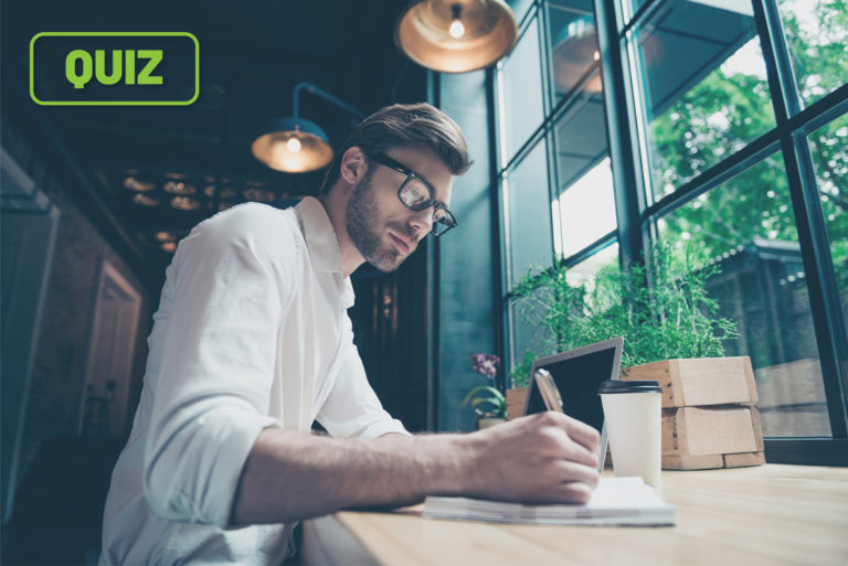 A young professional sits at a table writing in his notepad next to his computer.