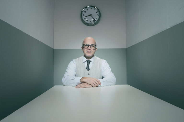 Intimidating hiring manager sits at the end of an interview table in a cold, outdated interview room