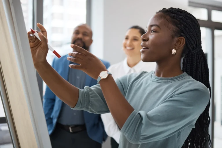 Woman using dry erase market to write on whiteboard with colleagues behind her.